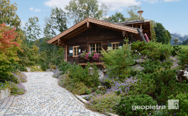 Questa immagine mostra uno chalet alpino tradizionale caratterizzato da un'armoniosa combinazione di legno e pietra ricostruita. Il tetto spiovente e il balcone decorato con fiori colorati evidenziano l'architettura rustica. Il verde della vegetazione circostante e gli elementi naturali come i muri in pietra ricostruita contribuiscono a creare un ambiente accogliente e pittoresco.